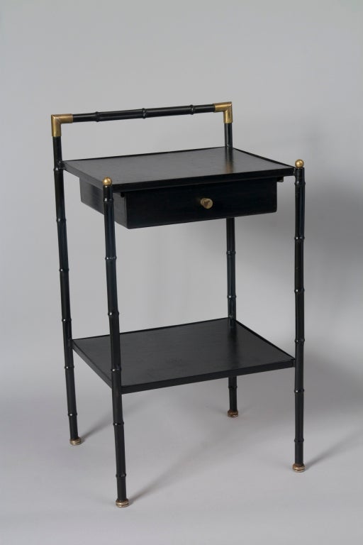 Faux bamboo frame two tier occasional table, in black lacquered metal with dore bronze caps and joints, black leather surfaces and a single drawer, by Jacques Adnet, French circa 1950. Overall height 26 1/2 in, height to top surface 23 in, width 14