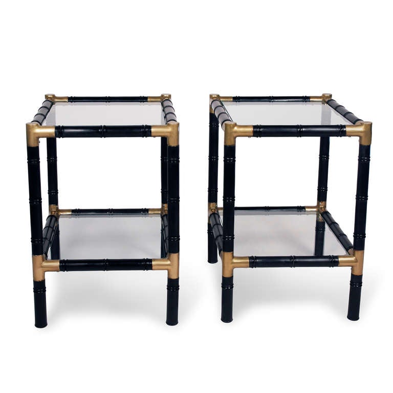 Pair of black lacquered faux bamboo wood two tier end tables, with bronze fittings on corners, and clear glass shelves. In the style of Billy Haines, American circa 1960. 19 1/2 in x 16 in, height 23 in. (Item #1308NB)