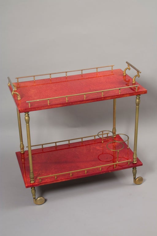 Two tier marbled dark red goat skin serving cart, with brass gallery edge, carved arced pull handles and wheel posts, the lower shelf having three bottle holders, by Aldo Tura, Italian, 1960s. 30 x 16 1/2 in, height to top surface 30 in. (Item #1539)