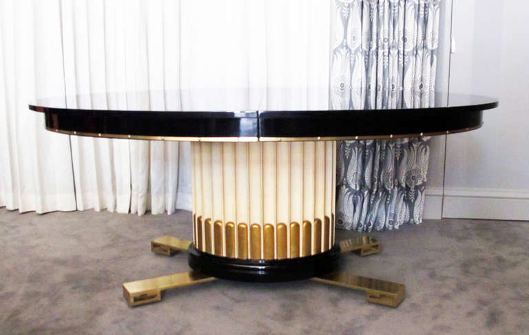 Here's a very decorative and well-crafted dining table possibly designed by Paul Frankl. The ivory lacquered and gold leafed column rest of four heavy brass feet. Table has two 14-inch leaves reflected in dimensions listed below. Black-lacquered top