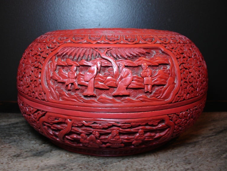 A large and finely carved Chinese cinnabar lacquer round, covered cake box. The body deeply caved with a chrysanthemum and scrolling foliate pattern. Eight different scenes of scholars with children in a garden setting are carved in the reserve