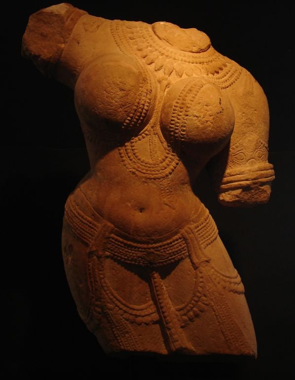 A wonderfully sensual torso of a female deity. 
Carved from indigenous pink sandstone, she is portrayed in an exaggerated tribhanga (thrice-bent) pose. She wears a diaphanous dhoti and is richly adorned. 
The low slung belt and jeweled festoons