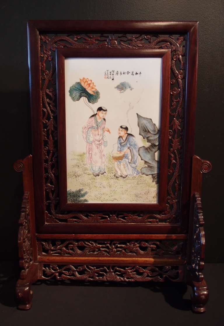 A famille rose enameled porcelain plaque featuring the Hehe Erxian (the immortal twin boys of Harmony and Union). The boys are portrayed seated in a garden setting dressed in informal court robes, one in blue, the other in pink, holding their