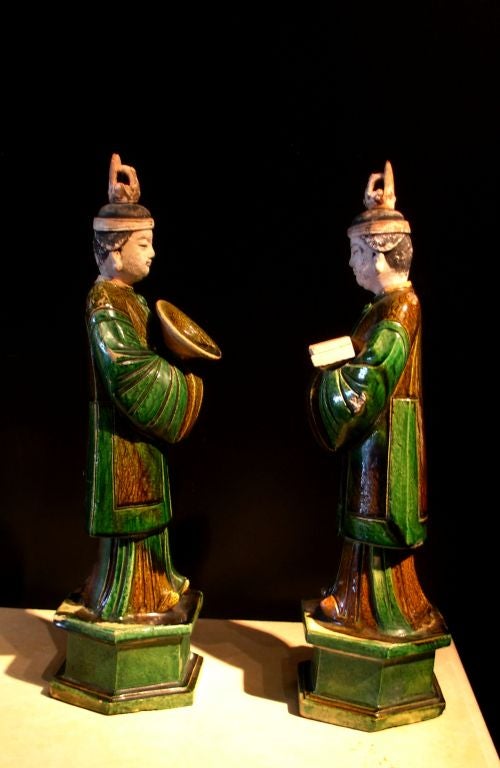A pair of well molded and glazed figures of attendants. Each portrayed standing on a hexagonal plinth and wearing long robes and a surcoat typical of the period, their hair neatly tied into a topknot and secured by a headband and cover. Both hold