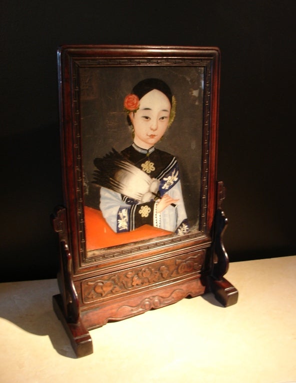 An unusual table screen featuring a reverse glass painted portrait of a young noble lady set into a hardwood frame and stand.
The beautiful maiden has a round face with a high forehead and almond shaped eyes. She is portrayed wearing an informal