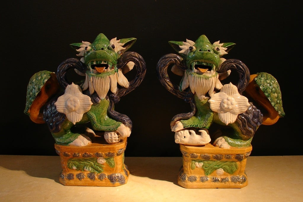 A charming and whimsical pair of Chinese glazed pottery foo dogs. 

Raised on rectangular plinths, each carries a streaming ribbon in their open mouth and are highly animated, with bulging eyes and lolling tongues. A large magnolia blossom