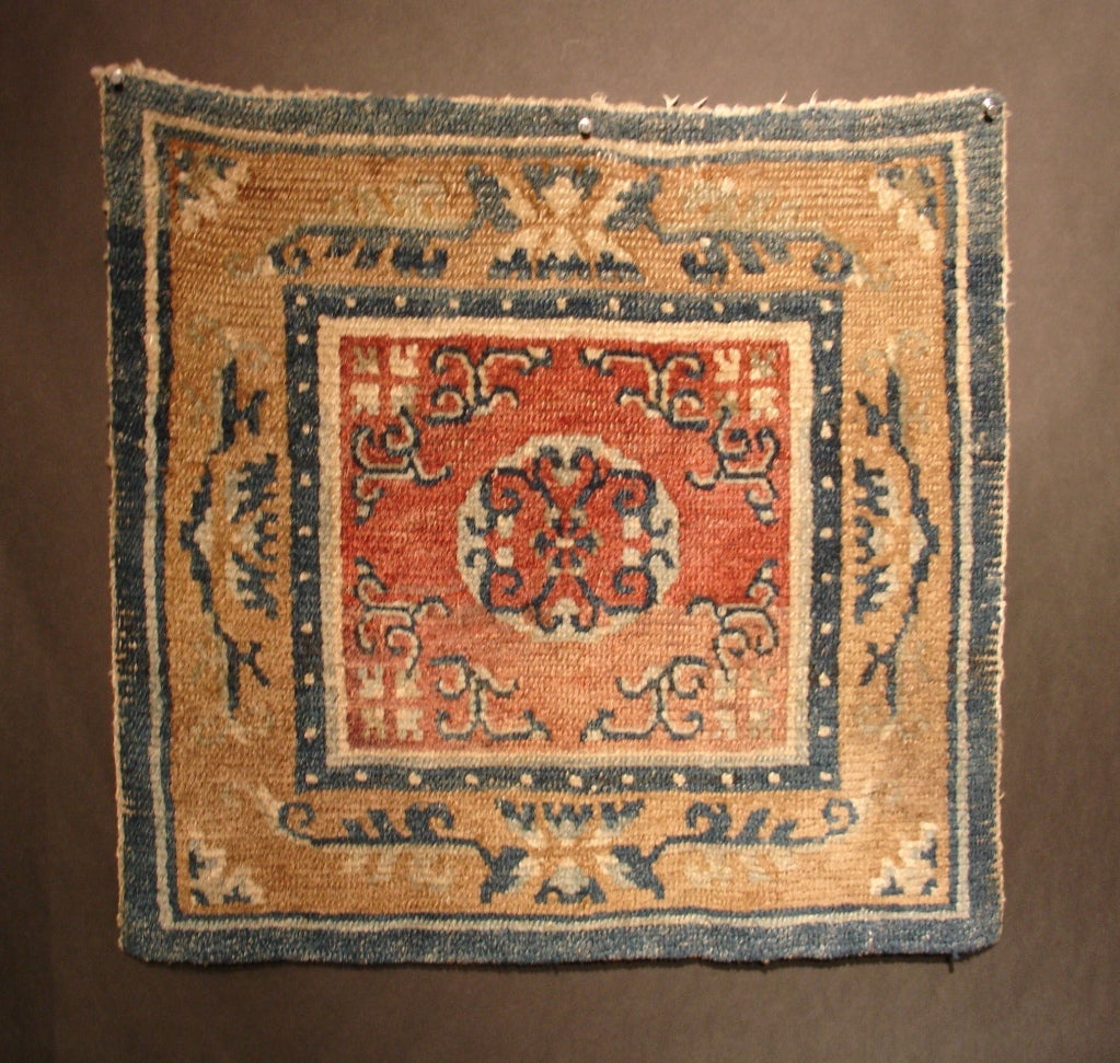 An early Tibetan wool square meditation mat featuring a central medallion on a red field. Eight stylized tigers form a lozenge shape around the central medallion. Blue lotuses on a yellow ground form the main border.