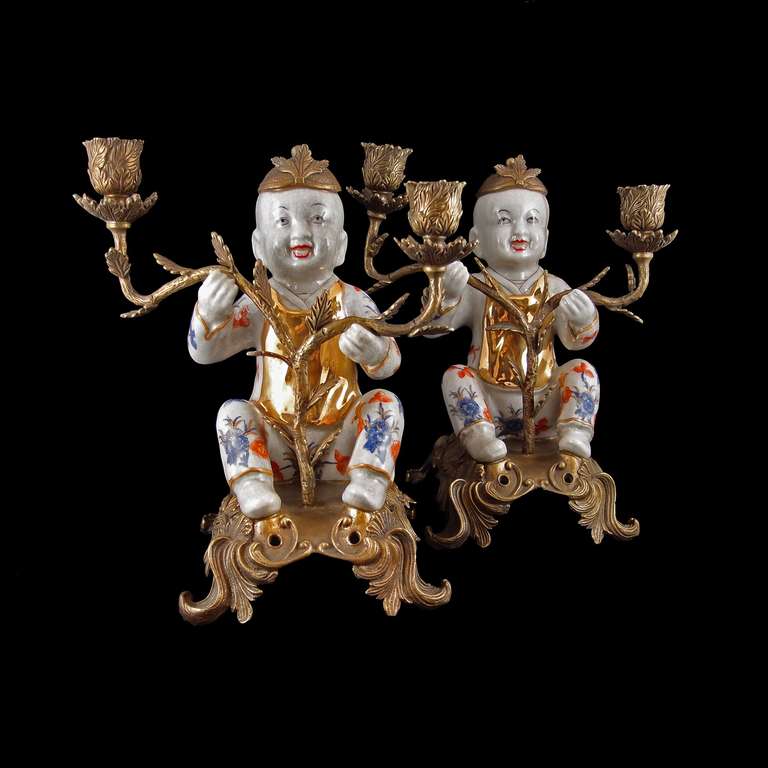 A delightful pair of two-arm candelabra, each featuring a porcelain figure of a laughing boy set in ormolu mounts. The boys, decorated in the Imari style, are posed in a seated position, arms outstretched, a joyous expression on their faces. A