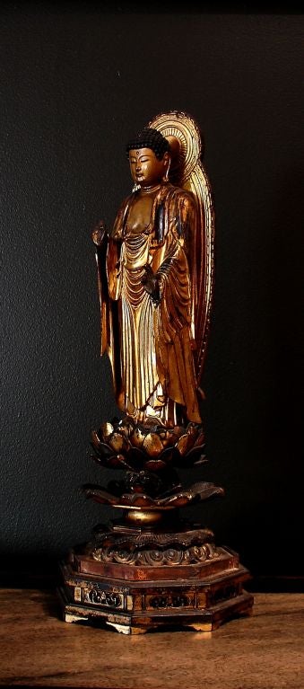Carved and richly gilt, this statue represents the historical Buddha, Shakyamuni. Standing on an open lotus blossom set upon an elaborately carved dais, he displays the abhaya mudra, the gesture of fearlessness and protection. He wears loosely