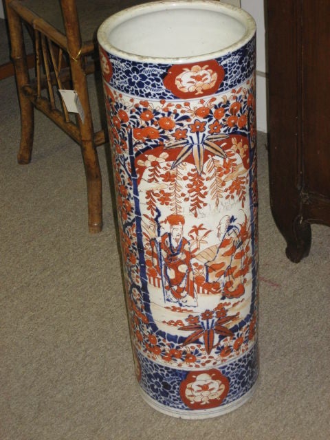 Pair of Japanese umbrella stand from the late 1800s. Stand is decorated with a center panel showcasing two men in a garden in traditional Japanese garments surrounded by a floral design. The piece has glazing in colors of blue and iron red with
