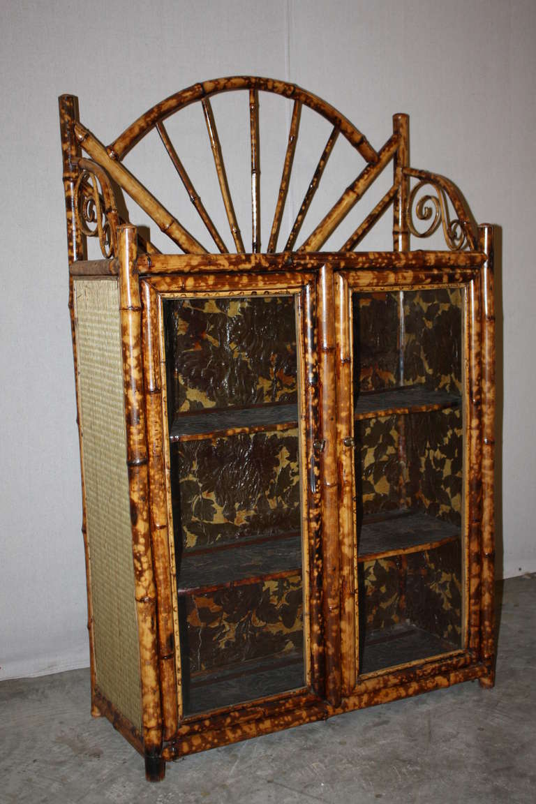 This is a very good looking small bamboo bookcase from the late 1800's.  It has a chinoiserie painted top.  It has two glass front doors.  The interior has three shelves.  The interior is made of a leather that is cut in the shape of leaves to