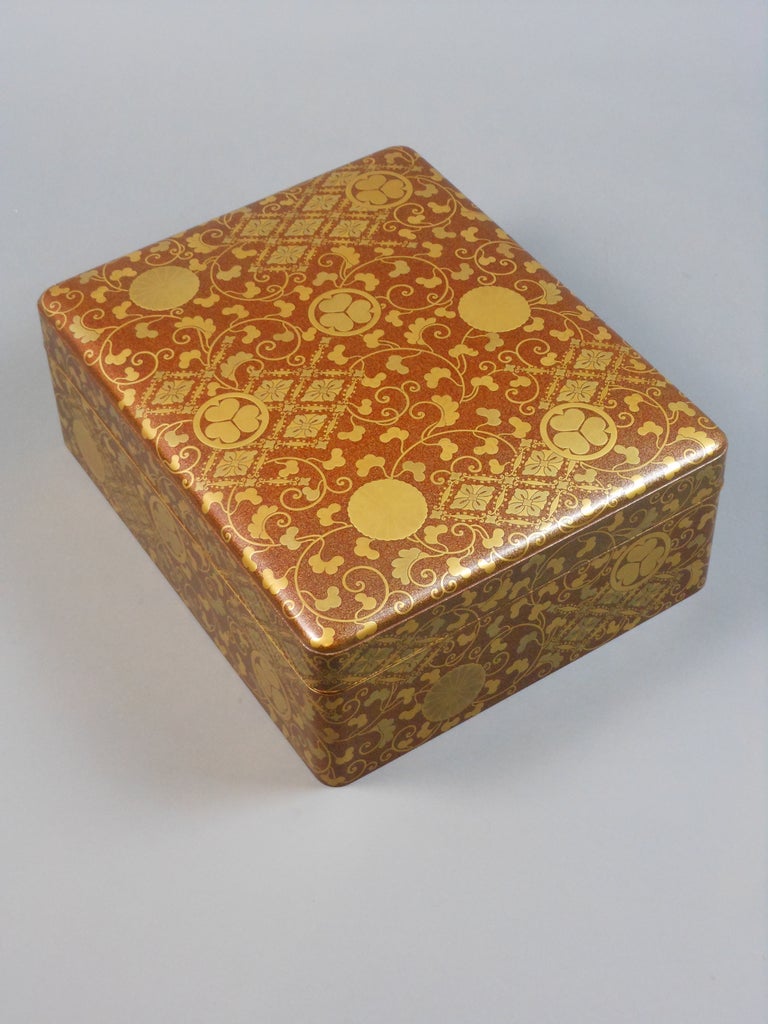 The rectangular lacquer box has rounded edges. The exterior depicts scrolling vines interspersed with the imperial kamon and the togukawa mon on a brown and gold maki-e background. On the underside of the cover, the spectacular low relief lacquer