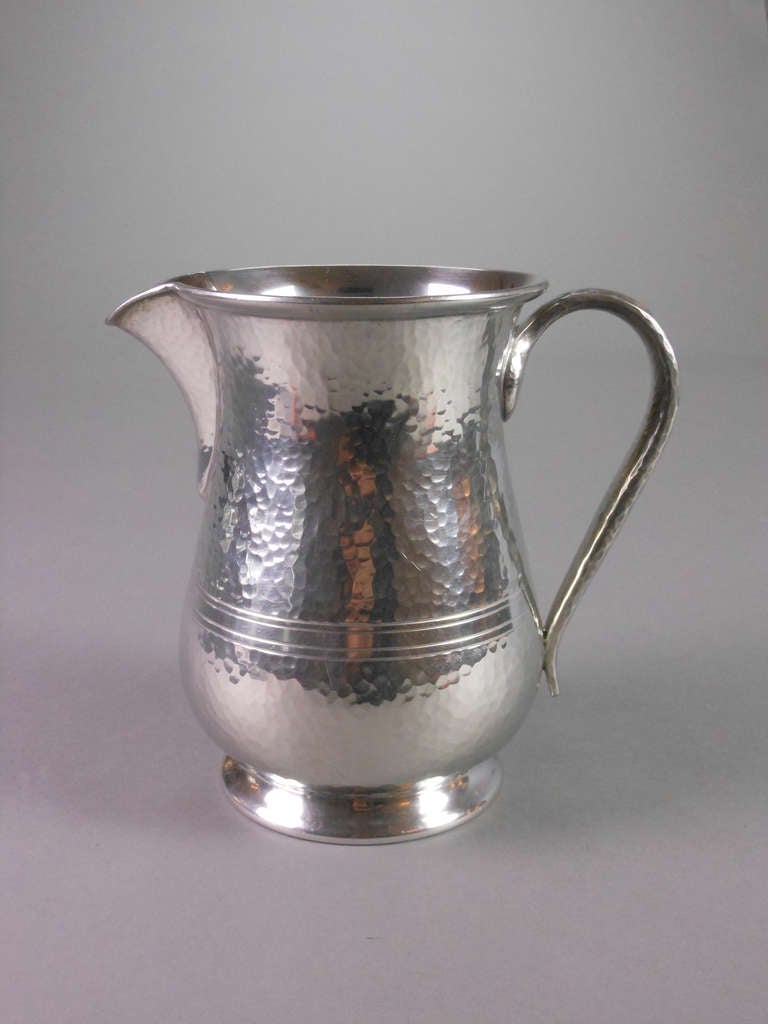 This English pewter jug has a hammered surface with a band of incised lines and an elegant scroll handle. By Archibald Knox for Liberty. Stamped "Made in England, BCM Tudric 01681, 5 pints."