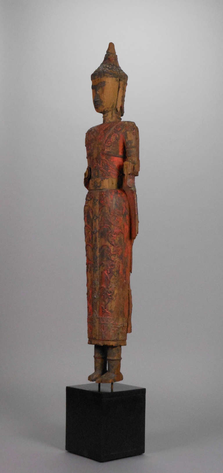 The Thai carved, elongated figure of the Buddha has a beautiful red and black patina surface. The scrolling relief on the Buddha's robe suggests elaborate embroidery. The standing Buddha faces forward; the missing forearms suggest the abhaya mudra