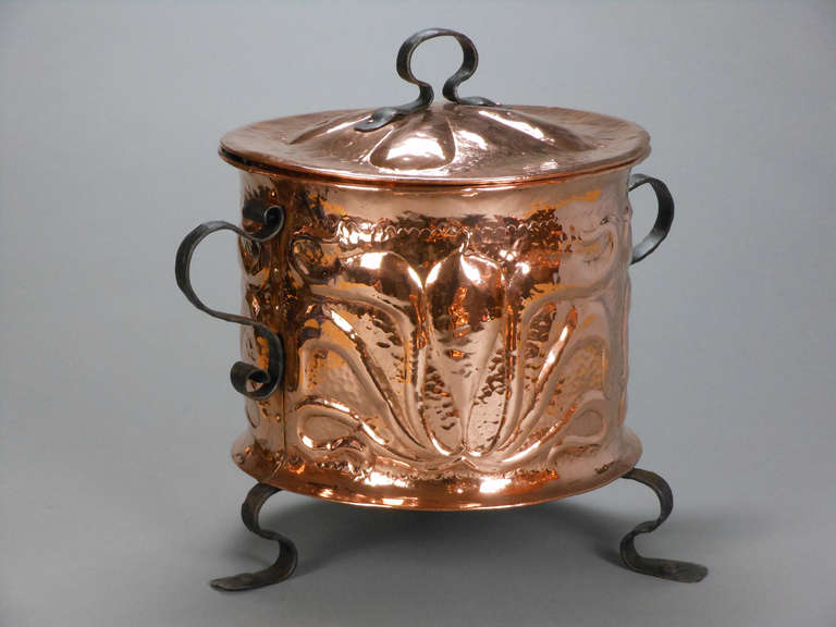 This Art Nouveau copper pot has a circular domed lid with a hoop handle. The cylinder body has a scrolling foliate repoussé decoration and is fitted with scroll handles and feet.