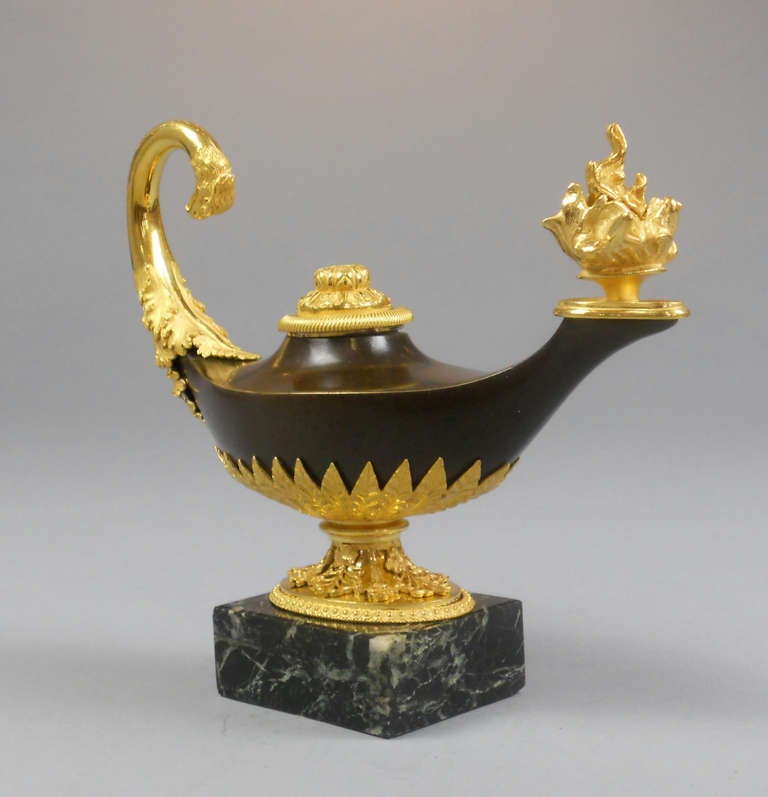 The candlestick is in the form of an oil lamp. The curving handle ends in an eagle's head and the spout issues a flame that is used to douse the candle. The rosette-mounted nozzle reverses to a candleholder. The lamp is mounted with a beautiful leaf