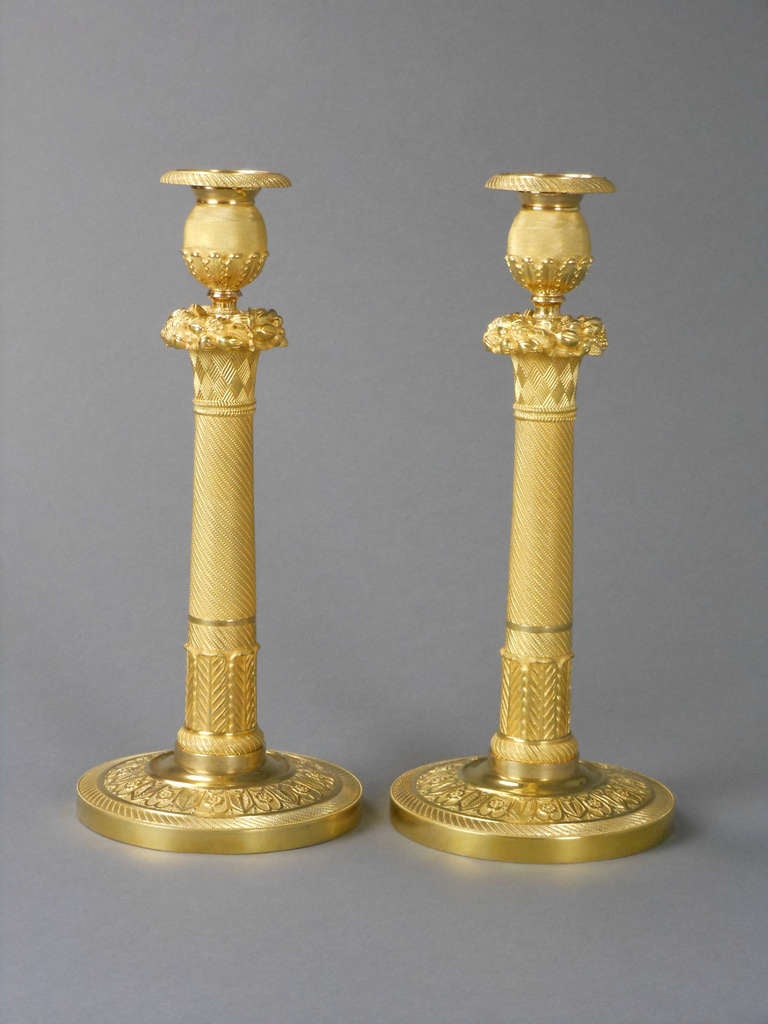 Each candlestick has a tapering, engine-turned stem with a cornucopia basket and an ovoid candleholder with acanthus leaf frieze. The domed base has a frieze of alternating rosettes and anthemion leaves.