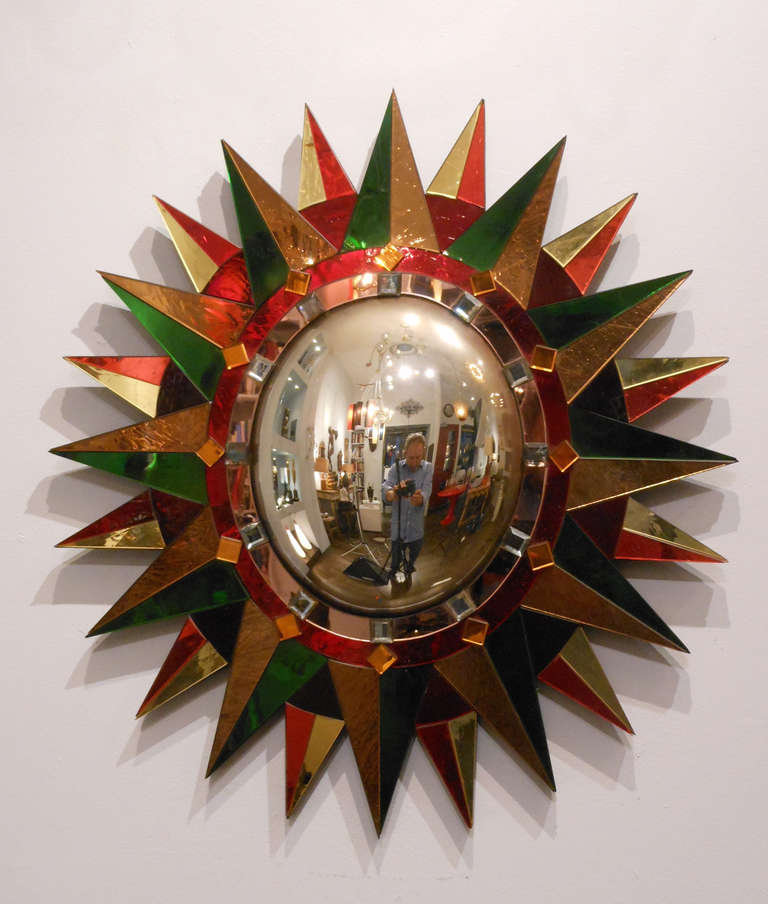 The oval convex mirror is framed by pink and red bands applied with faceted squares, in turn surrounded by green, red and yellow overlapping rays.