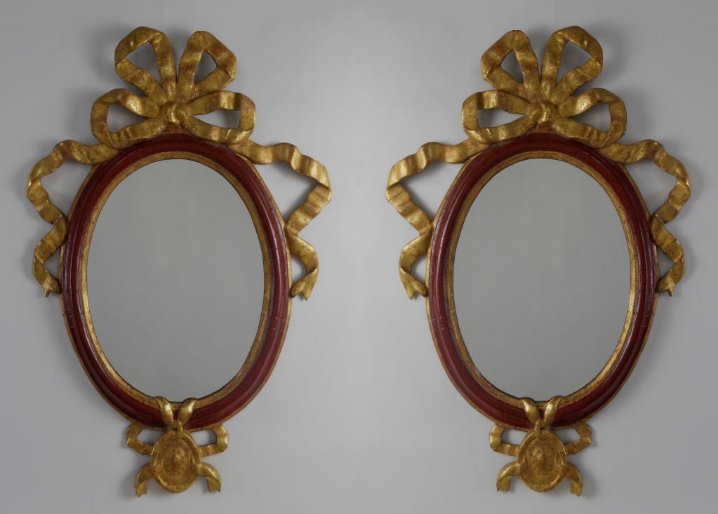 The neoclassical mirrors have an oval mirror plate within a gilt and red lacquered molded frame mounted with a bow and trailing ribbons.