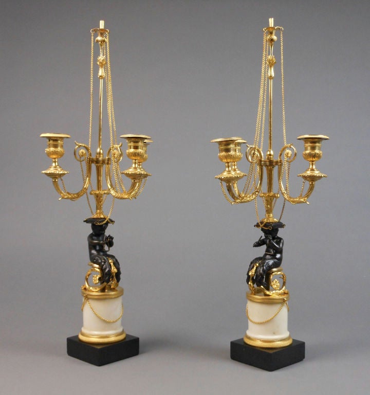 Each of these French neoclassical candelabra has a putti triton seated on a sleigh playing a triangle. The foliate standard issues three scroll patera arms ending with urn form candleholders and linked with chains. The circular white marble column