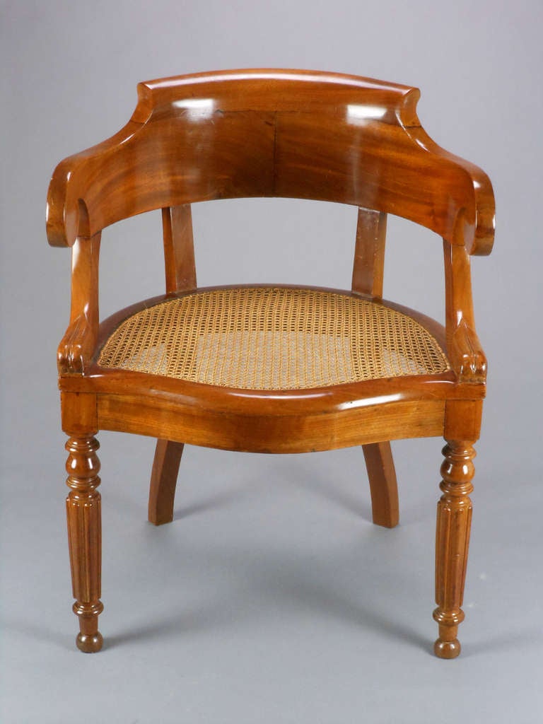 The Charles X period chair has a flared top rail above the curved backrest and scroll arms on downswept supports. The caned, shield form seat is raised on round tapering fluted front legs and saber back legs.