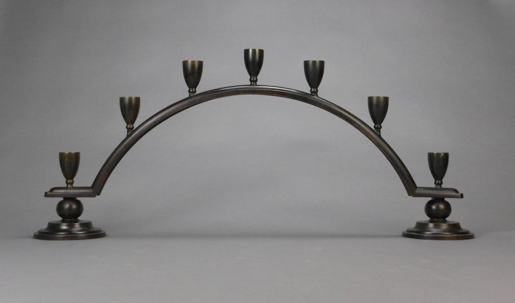 The candelabra is a molded arch fitted with seven urn form candle holders, each end raised on a sphere and stepped circular base. Stamped DENMARK JUST A, B173.

Just Andersen (1884-1943), the celebrated Danish bronzier, created outstanding patinated