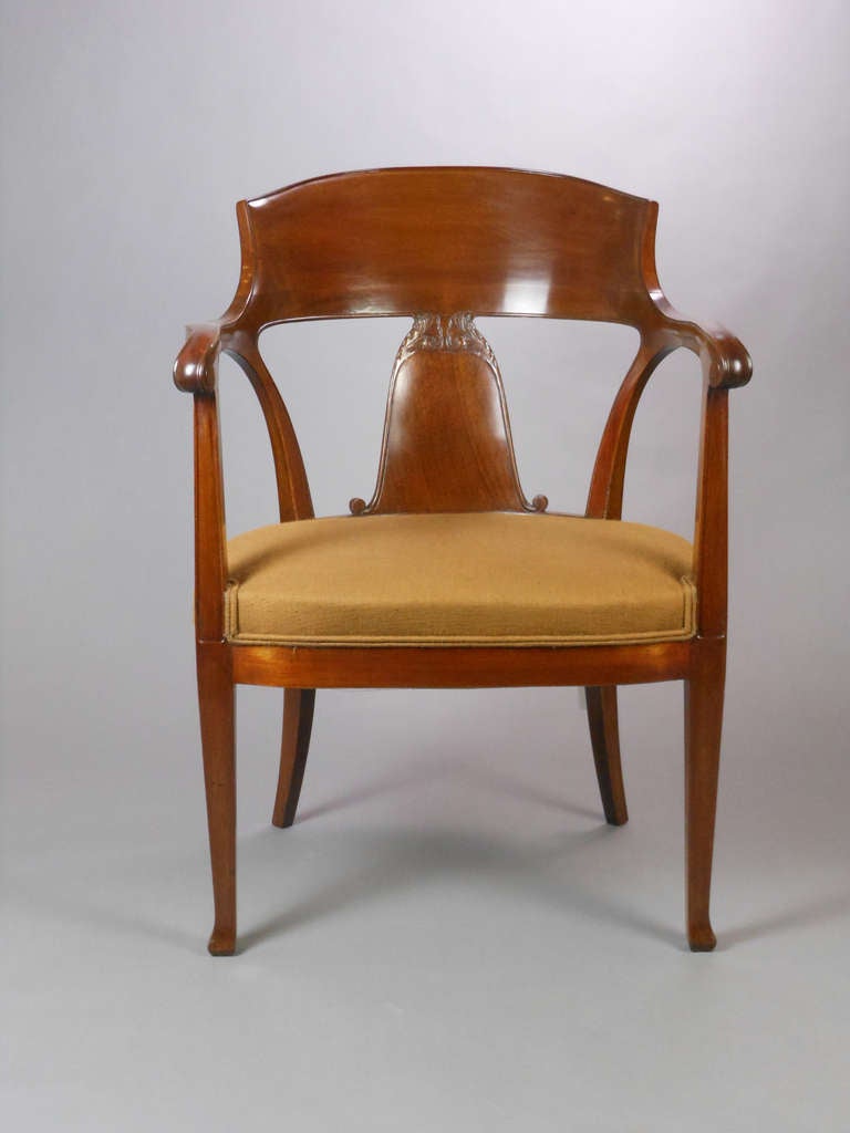 This graceful mahogany wood chair has a curved backrest and a bellflower-shaped splat with low relief foliate scrolls. The chair features scrolled armrests above and square tapering legs below the upholstered seat.