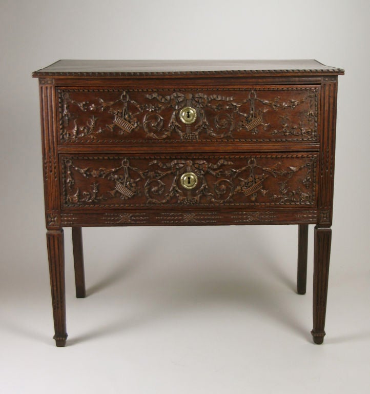 This richly carved Italian mahogany commode has a molded edge ribbon twist top above two drawers. Each drawer is centered by a tied bow issuing scrolling bellflowers hung with baskets of flowers and musical instruments, all within a ribbon twist