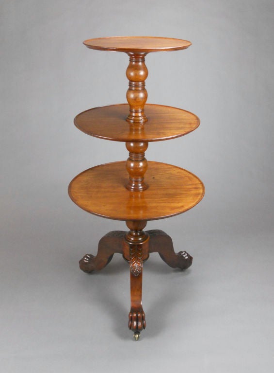 This French three-tiered table has a banded baluster standard that supports three graduating circular shelves with molded rims. The scrolled tripod base ends in paw feet on casters.