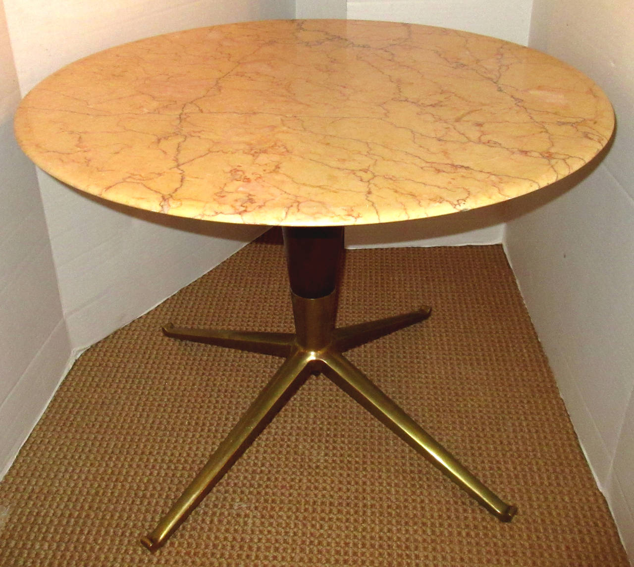 A low, round, honed marble topped end or side table. A tapered wood pedestal base stands on four solid cast brass splayed legs. The marble is heavily veined in tones of cream, copper and gray.