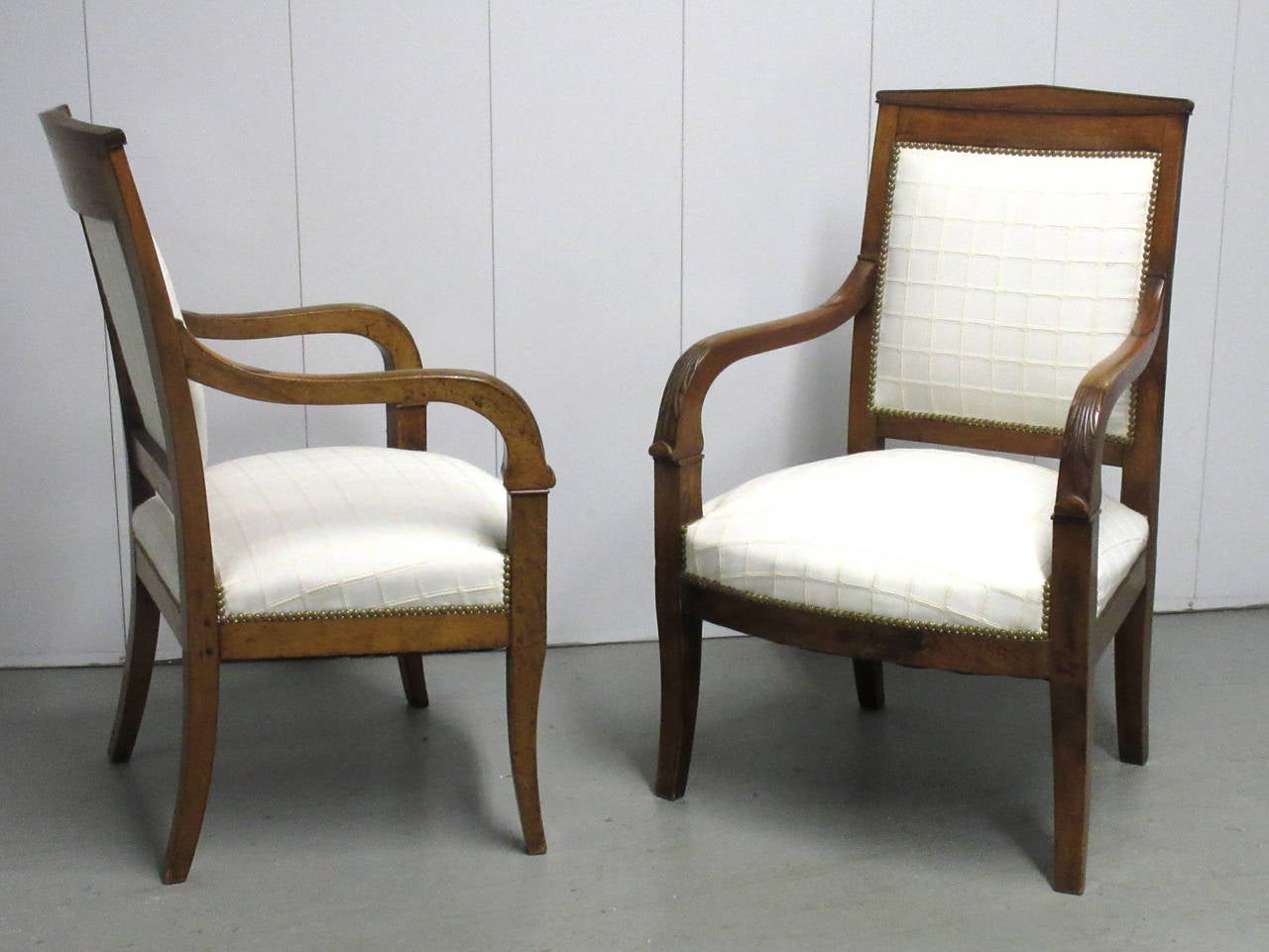 A pair of French Empire period upholstered armchairs with saber legs and foliate carved armrests.
Upholstery is new and embellished with brass nailheads throughout.