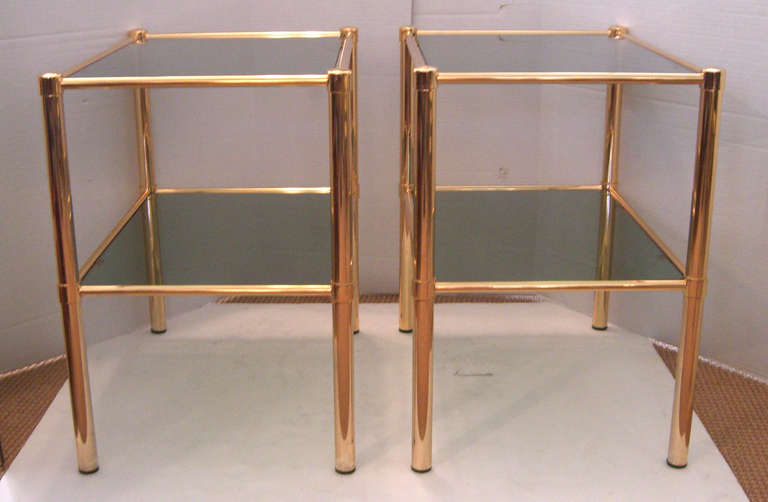 A pair of rectangular end tables in a high shine gold tone. The top and lower shelf are a gray smoked glass.