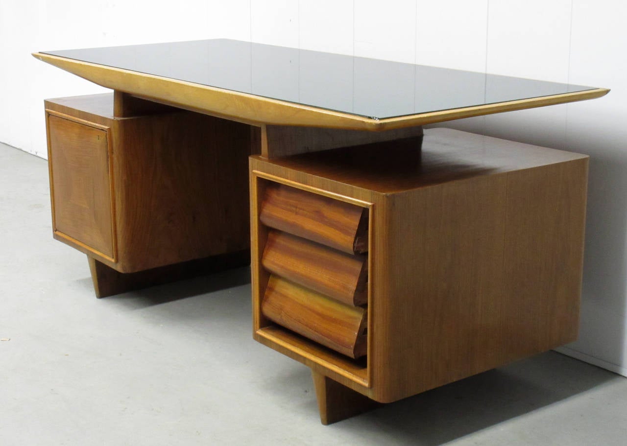 A large 1950's era Italian executive desk with a floating top. The top is inset with a dark green glass panel. The right side features 3 drawers with satinwood veneered fronts. The left side is a square cabinet space.