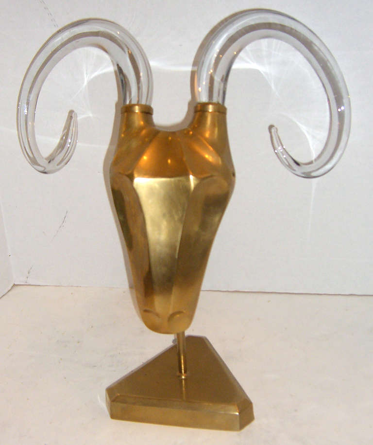 A stylized solid brass ram's head in a stand with large curving clear glass horns.