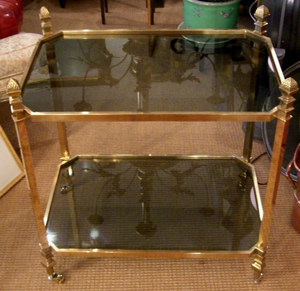 Beautiful gold tone brass table with four large pinecone finials on casters. Two shelves of gray toned glass.