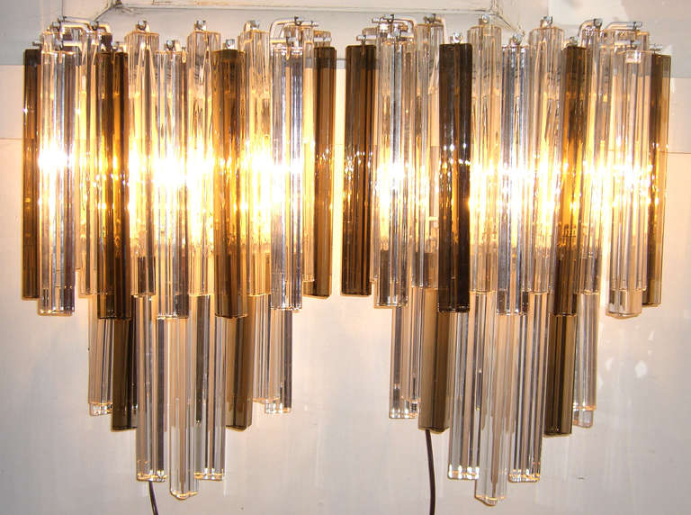 Grandi prismi in vetro Venini in una combinazione di trasparente e fumé. I prismi triedri in vetro si alternano in altezza per creare un effetto a cascata. Le applique sono illuminate da tre prese a candelabro appena cablate. È stata aggiunta una