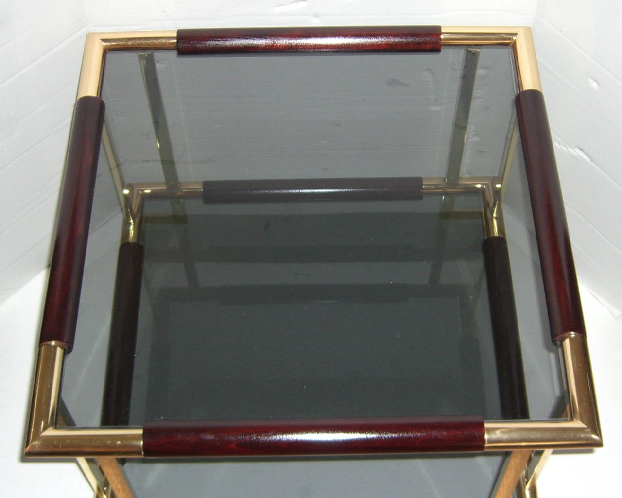 A square side table with a polished brass and dark walnut toned wood frame. The upper and lower glass shelves are smokey grey.