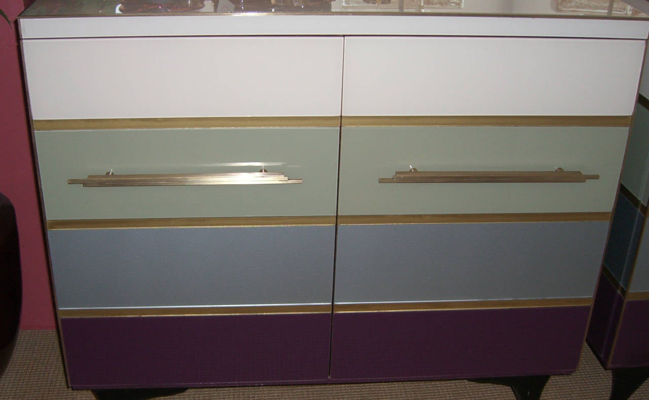 A pair of two door cabinets with panels of back painted glass. The panels are in beautiful tones of plum, pastel blue, green and ivory. The trim and door pulls are brass, and the legs are ebonized wood. The inner cabinets are high shine lacquered