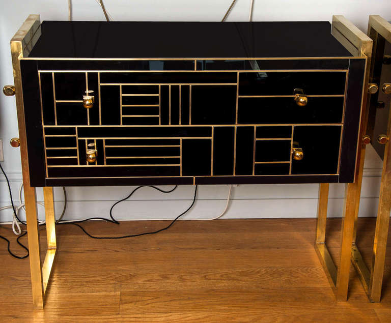 Stylish pair of Italian 1950's black glass and brass chests each with two drawers. Each drawer is inlaid with slim geometric brass lines and features elegant brass sculptured handles. Stands on a simple brass frame/legs with four further sculptured