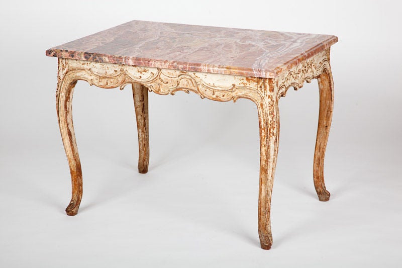 This mid-18th century French Louis XV period centre table has cabriole legs and asymmetrical rocaille carving, with cream painted wooden surface, beneath marble top.