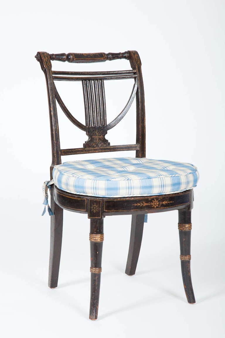 This early 19th century English Regency period side chair has a lyre form open work back above a round caned seat, raised on splayed legs, all retaining its original parcel-gilded decoration on a painted black ground.