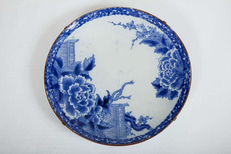 This large Japanese Meiji period porcelain igezara charger is decorated with large scaled peonies, cherry blossoms and bamboo fences in blue on a white ground, and now mounted to brass strap for hanging. Igezara ware was heavy, durable porcelain