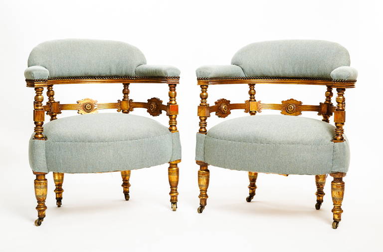 This pair of English Victorian period tub chairs have upholstered upper back and arm rests above gilded turned wooden arm and back supports joined by stretchers with stylized flower heads, connecting to an upholstered seat, raised on turned tapered