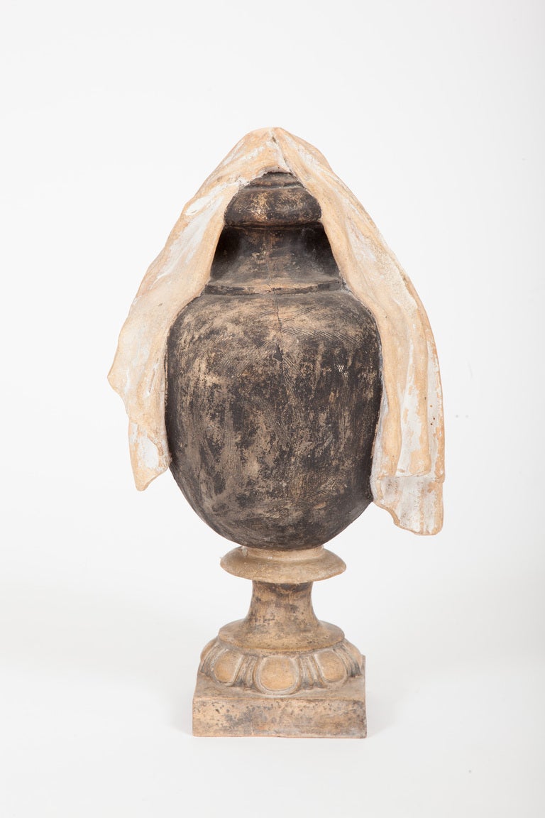 This pair of Italian neoclassical urns is made of painted terra cotta, each with a modeled piece of fabric draped over a lidded ovoid shaped urn, the body painted black, raised on a socle with egg and dart molding, supported by a square plinth