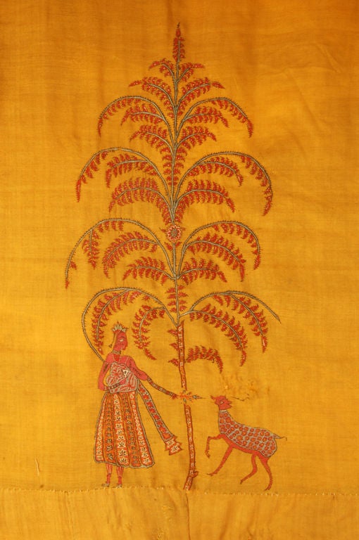 Large temple hanging of saffron yellow featuring Krishna and Rhada, each playing with a deer under a flowering tree in the center of a plain oval field that is surrounded by borders worked with flowers, botehs and large birds in corners.
