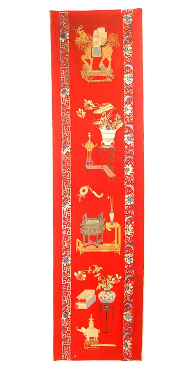 A long and narrow wall hanging of red wool decorated with a column of embroidered subjects. The top is the mythical animal xilin and the bottom is an elephant. In between them are articles of an interior with furniture and vases with flowers. The