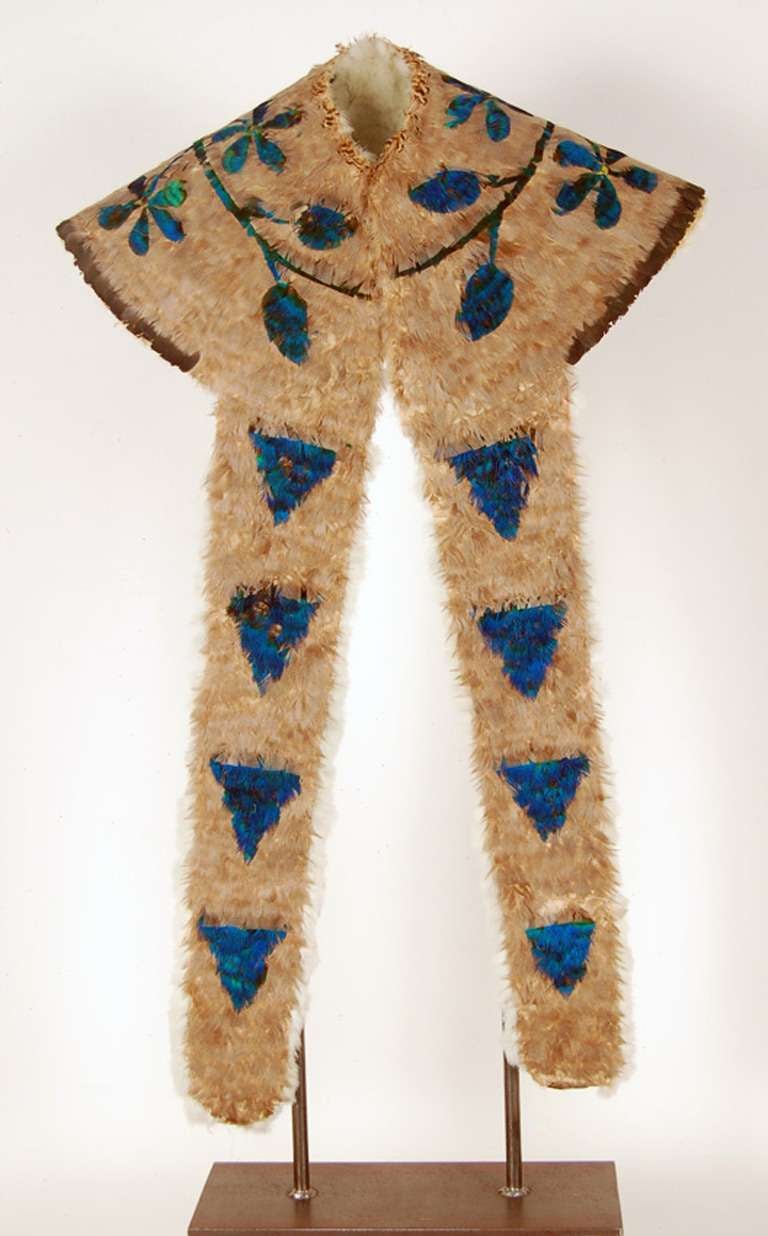 Circular cape with tapered long ends. A design two flowering sprigs that originate at the back is on the round top, while the two front ends feature triangular shapes. All done with blue green iridescent feathers against a light brown small turkey