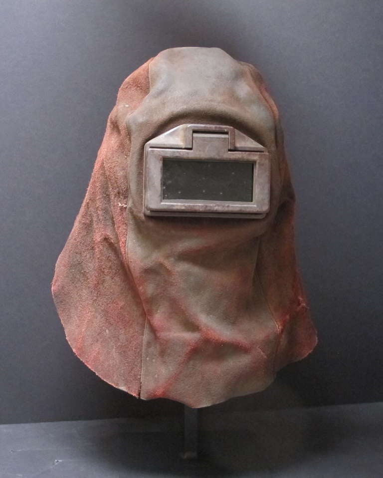 Protective leather helmet mask with dark glass eye window. It has an adjustable tie in the back and small upper vents. Unusual colored leather with patina from use. A mask for protection mounted over a black metal base.