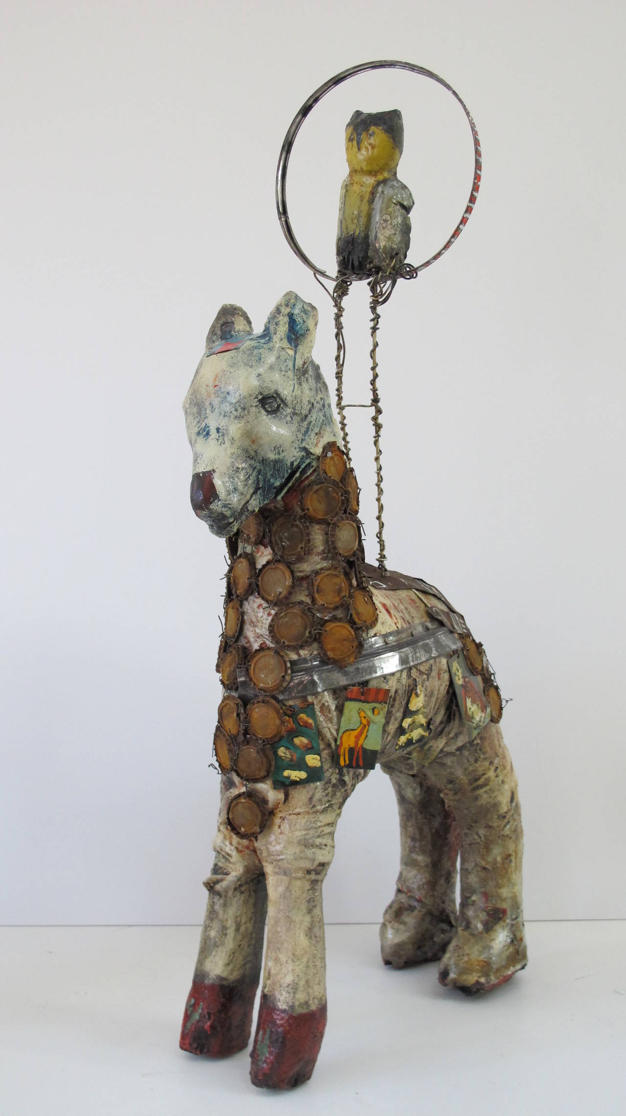 Sculpture by self-taught artist Terry Turrell of carved wood horse with an owl perched above in a ring. The sculpture is painted and has applied pieces of cut tin and is draped with vintage bottle caps wired together.