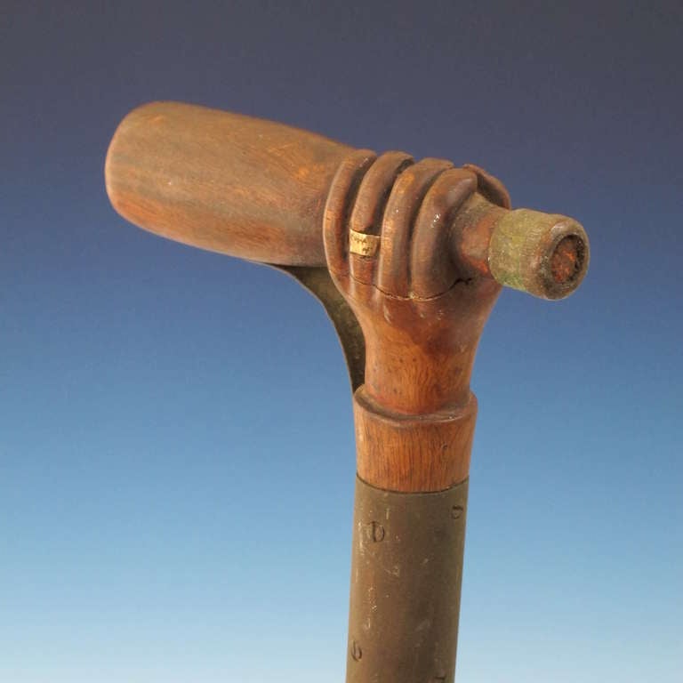 Well carved wood hand with metal ring on finger grasping a bottle with copper top cap.  The knobby wood cane becomes the arm with a metal cuff and an arching brace added to the bottle for strength.  The bottom has a brass ribbed ferrule. Mounted on